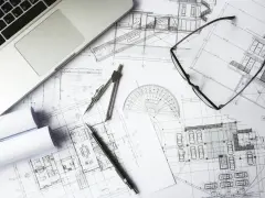 Na biurku architekta leżą plany budowlane, laptop, okulary i cyrkiel. Trwają prace nad przedmiarem robót.
