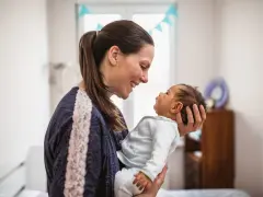 Uśmiechnięta mama trzyma na rękach noworodka. Dziecko patrzy na nią z zaciekawieniem, jakby miało zaraz kichnąć.