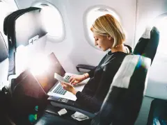 Kobieta pracuje na laptopie i trzyma telefon, zastanawiając się, czy w samolocie można ładować telefon.