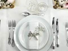 Ett elegant dukat bord med vita tallrikar, servetter dekorerade med blommor och vackra, silverfärgade engångsbestick.