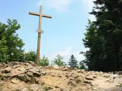 Drewniany krzyż na skalistym szczycie, najwyższy szczyt w górach świętokrzyskich, otoczony zielenią drzew.