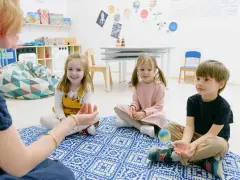 Dzieci siedzą na dywanie, słuchając nauczycielki. To przykład metody dobrego startu, gdzie nauka odbywa się przez zabawę.