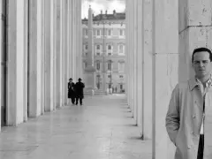 Un uomo in trench osserva l'obiettivo, mentre sullo sfondo due figure in abito scuro passeggiano sotto un colonnato. Potrebbe essere una scena da "Il talento di Mr. Ripley dove è stato girato".