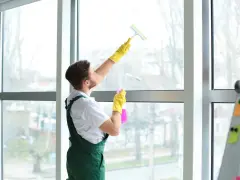 Mężczyzna w żółtych rękawiczkach i zielonym ogrodniczkach pokazuje, jak wyczyścić uszczelki w oknach, używając sprayu i ściągaczki.