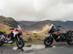 Due moto, una Ducati Multistrada 1290 rossa e bianca e una KTM 1290 Super Adventure nera e arancione, parcheggiate su una strada di montagna con un paesaggio nebbioso sullo sfondo.