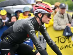 Trine Vingegaard, a cyclist in a grey and black racing suit, rides in the rain. Spectators watch from behind a yellow barrier with "Tour de France" branding.