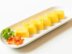 Sześć kawałków puszystego omletu tamago ułożonych na białej tacy, ozdobionej natką pietruszki i pomarańczowym dodatkiem.