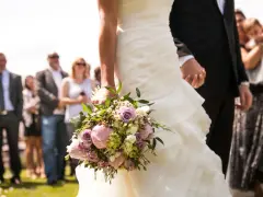 Panna młoda trzyma bukiet, a jej partner na wesele trzyma ją za rękę. W tle widać gości.