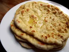 Stos świeżo upieczonych, lekko przypieczonych placków roti i naan, polanych roztopionym masłem, na białym talerzu.