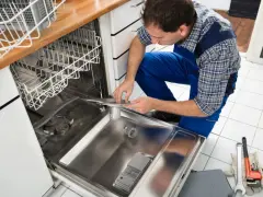Fachowiec w niebieskim kombinezonie montuje element zmywarki, pokazując, jak podłączyć zmywarkę. Obok leżą klucze i rękawice.