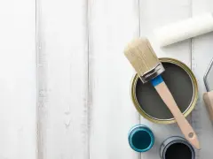 Herramientas para pintar mesitas de noche: lata de pintura gris, pincel, rodillo y botes de pintura azul y negra sobre fondo de madera blanca.