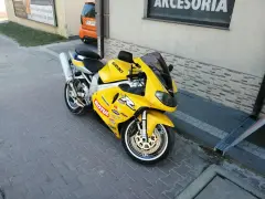 Żółty motocykl Suzuki TL1000R z naklejkami sponsorów, zaparkowany na chodniku.