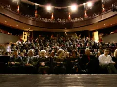 Widownia teatru, ludzie siedzą na miejscach, widać balkon z widzami.