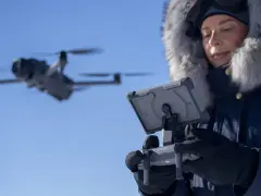 Woman in a fur-lined hood controls a flying drone in cold weather, looking at a tablet.