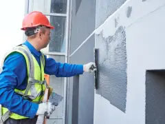 Pracownik w kasku i kamizelce nakłada tynk na ścianę budynku, prezentując różne rodzaje tynków zewnętrznych.