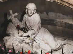 A stone sculpture depicts the serene body of Mary Magdalene, resting with a cross and a single red rose.