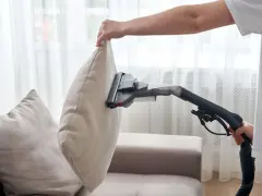 En person använder en soffdammsugare för att rengöra en ljusbeige kudde på en soffa.