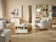 Jasny salon z sofą, fotelem i stolikiem kawowym na kółkach. Na półkach regału stoją dekoracje, książki i lampa. Wersaliki dodają elegancji.