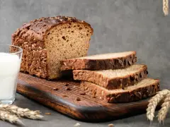Chleb proteinowy z ziarnami, pokrojony na kromki, obok szklanka mleka i kłosy zboża. Idealny na zdrowe śniadanie.