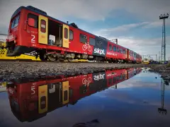 Czerwony pociąg SKPL, stowarzyszenie kolejowych przewozów lokalnych, odbija się w kałuży. Obok żółty tramwaj Kolei Dolnośląskich.