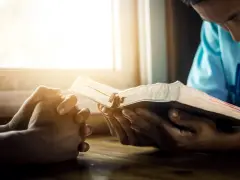 Hands clasped in prayer beside an open Bible, bathed in warm light. A moment of reflection, perhaps contemplating prayer names.