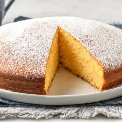 Torta yogurt greco soffice e dorata, cosparsa di zucchero a velo. Una fetta è stata tolta, rivelando l'interno profumato.