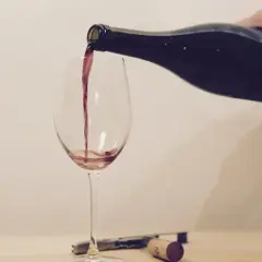 Ein Weinglas wird mit Rotwein gefüllt. Eine Hand hält die dunkle Flasche, während der Wein in einem dünnen Strahl ins Glas fließt.