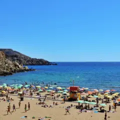 Słoneczna plaża na Malcie, idealna na wycieczkę na Malte last minute. Tłumy ludzi, kolorowe parasole i turkusowe morze.