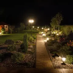 Piękna ścieżka z kostki brukowej prowadzi przez ogród, który nocą rozświetla nastrojowe oświetlenie. Inspiracje dla Twojego ogrodu.