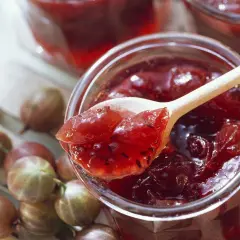 Ein Holzlöffel mit leckerer Stachelbeermarmelade ragt aus einem Glas. Frische Stachelbeeren liegen daneben.