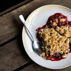Ein Teller mit warmem Pflaumen Crumble, dazu ein Löffel auf rustikalen Holzdielen.