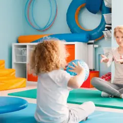 Dziecko z kręconymi włosami bawi się piłką, a kobieta instruuje je w sali do terapii SI.