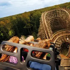 Fahrt mit einer Holzachterbahn, Menschen mit wehenden Haaren, im Hintergrund Wald und Himmel.