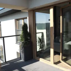 Nowoczesne okna dzielone z drewnianymi ramami otwierają widok na balkon z roślinnością i wnętrze domu.