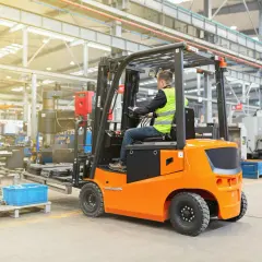 Ein Mann in Warnweste fährt einen orangefarbenen Gabelstapler in einer Fabrikhalle. Die Nutzungsdauer des Gabelstaplers ist entscheidend für die Effizienz.