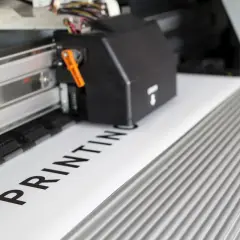 Drukarka wielkoformatowa drukuje napis "PRINTING" na białym papierze.