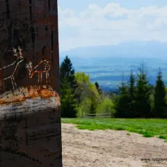 Rdzawy słup z rysunkami jelonków, symbolizujący najkrótszy szlak na Turbacz, na tle górskiego krajobrazu.