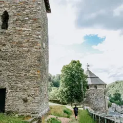 Ein Mann spaziert auf einem Weg zur Burg Monschau, vorbei an alten Steintürmen und grünen Hügeln.
