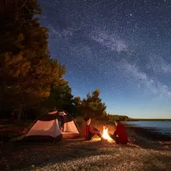 Nocne biwakowanie pod rozgwieżdżonym niebem. Dwoje ludzi siedzi przy ognisku obok namiotu z materiału namiotowego, na kamienistej plaży.