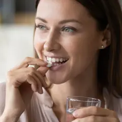 Femme souriante prenant un comprimé blanc, rappelant l'importance de ne pas associer le metronidazole alcool. Verre d'eau à la main.