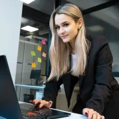 Młoda kobieta w czarnej marynarce pochyla się nad laptopem, pracując nad projektem. Jej studia biznesowe nabierają tempa.