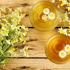 Due tazze di tisana dorata con fiori di camomilla galleggianti, accanto a un mazzo di fiori freschi. Rimedio naturale per il bruciore di stomaco.