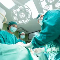 Zespół chirurgów w zielonych fartuchach i maskach przeprowadza operację nosa pod jasnym światłem lampy.