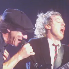 Dwóch członków AC/DC na scenie: wokalista z mikrofonem i gitarzysta w garniturze.