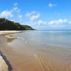 En stilla stenshuvud strand med klart vatten som möter en sandig kustlinje. Grönska och kullar syns i bakgrunden under en blå himmel med vita moln.