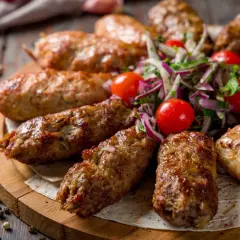 Kebab ormiański na drewnianej desce, podany z sałatką z pomidorków i cebuli.