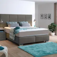 Ein modernes Schlafzimmer mit einem grauen Boxspringbett, das mit einer geometrisch gemusterten Decke und Kissen bezogen ist. Ein türkiser Teppich liegt davor.