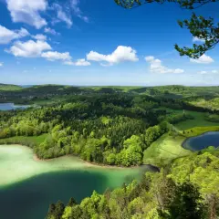 Paysage verdoyant du Jura avec des lacs aux eaux claires, des forêts denses et un ciel bleu parsemé de nuages blancs.