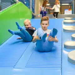 Dwóch chłopców zjeżdża po niebieskiej zjeżdżalni w sali zabaw. W tle widać inne dzieci. To świetna zabawa dla każdego dziecka!
