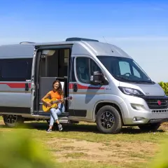 Une jeune femme joue de la guitare assise à l'entrée d'un camping-car aménagé, un type de camping car idéal pour les escapades ensoleillées.